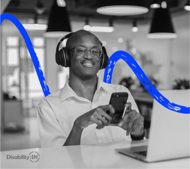 A person wearing headphones smiles while holding a phone, sitting at a desk with a laptop open.